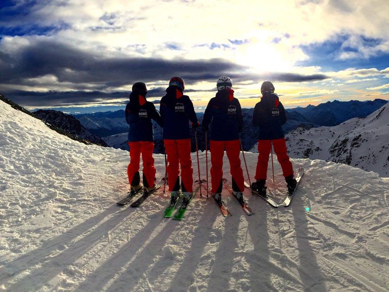 Cinque persone in controluce sulla pista, con le ombre lunghe sulla neve